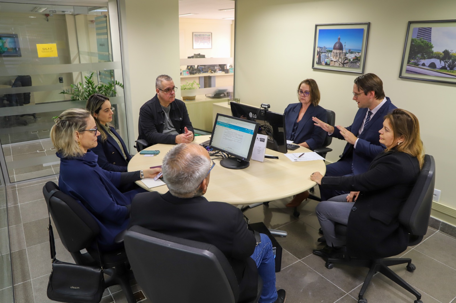 A foto mostra uma audiência de conciliação presencial, com pessoas ao redor de uma mesa.