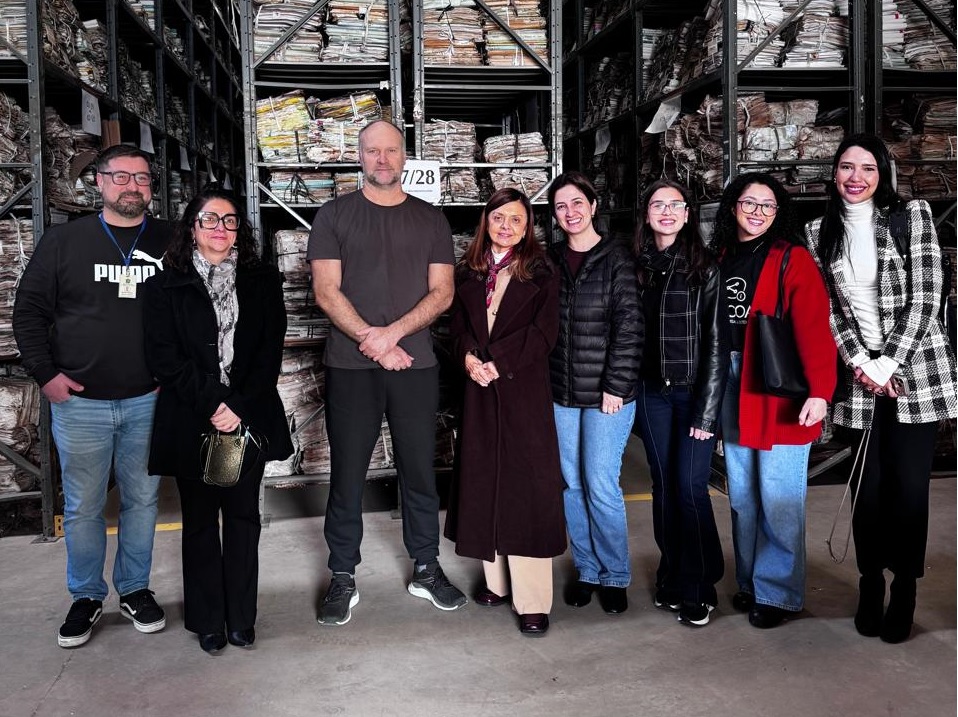 A fotografia mostra participantes da visita no Arquivo-Geral posando, lado a lado, em frente às estantes de processos do arquivo.