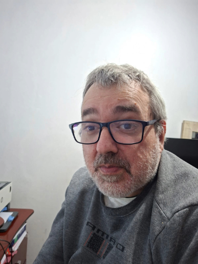 Márcio é um homem de pele clara, com barba e cabelos grisalhos. Ele está sentado à frande uma mesa na qual pode-se ver uma impressora. Usa moletom cinza e óculos de grau com aros escuros.