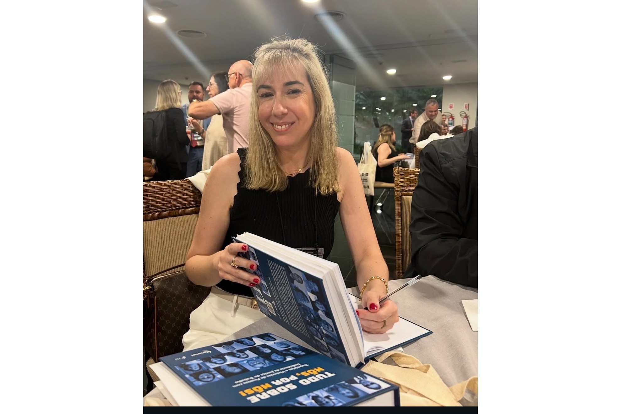 Renata é uma mulher de 40 anos. Ela tem a pele clara, cabelos loiros e olhos castanhos. Ela usa uma regata preta e calça bege. Está sorrindo, olhando para a câmera e autografa um livro. Atrás dela, há várias pessoas em pé.