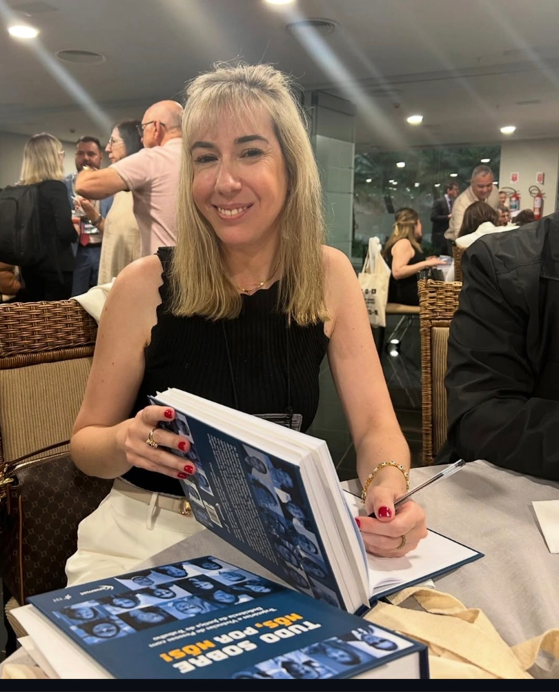 Renata é uma mulher de 40 anos. Ela tem a pele clara, cabelos loiros e olhos castanhos. Ela usa uma regata preta e calça bege. Está sorrindo, olhando para a câmera e autografa um livro. Atrás dela, há várias pessoas em pé.