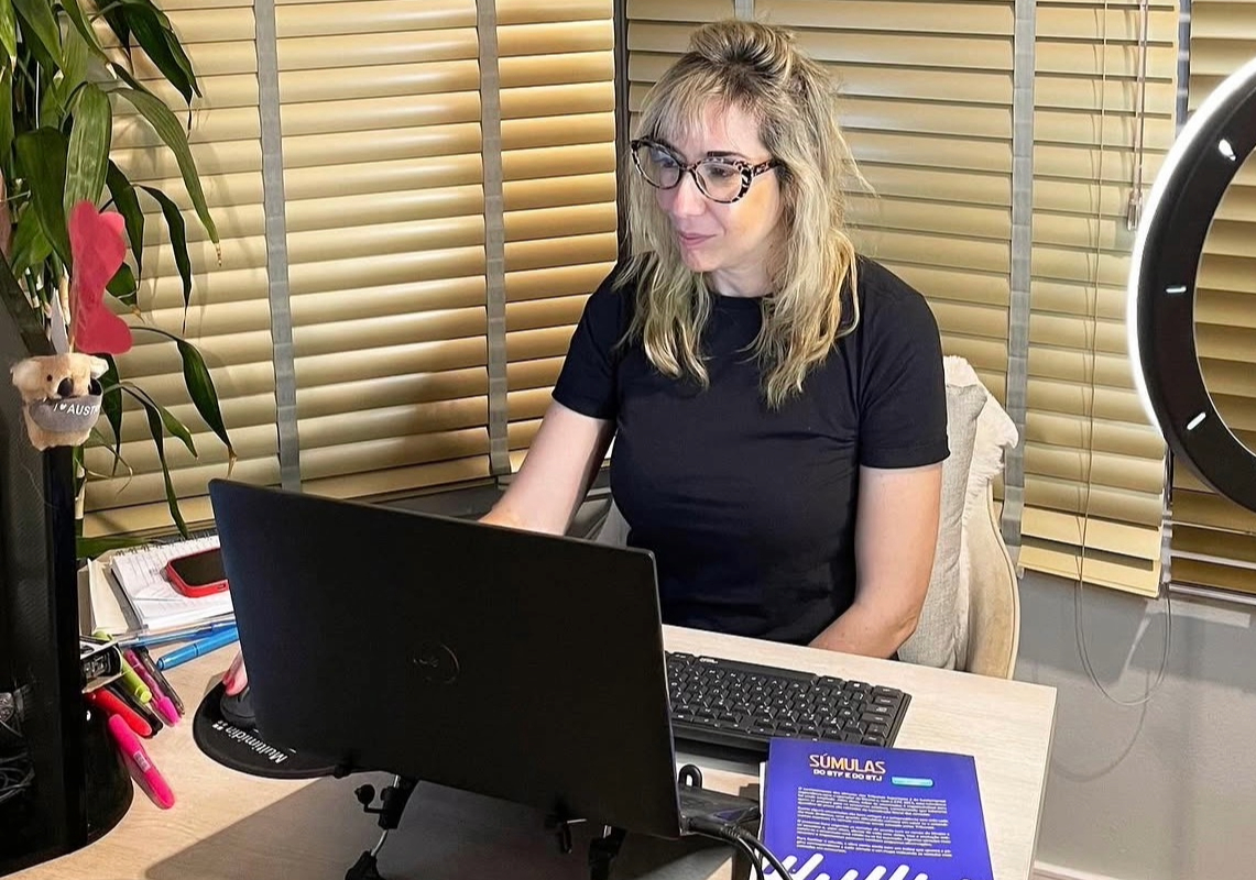 Renata é uma mulher de 40 anos. Ela tem a pele clara, cabelos loiros e olhos castanhos. Ela está em seu ambiente de trabalho, sentada em frente ao computador e com uma CLT sobre a mesa.