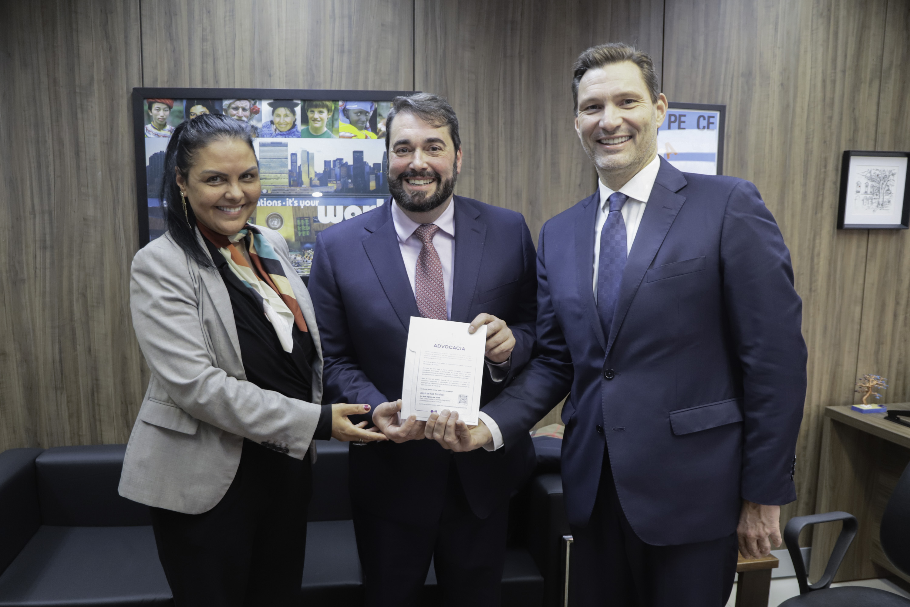 Tânia está na esquerda, Alexandre no centro e Leonardo na direita. Os três estão de pé, sorrindo, e seguram o convite para o evento.