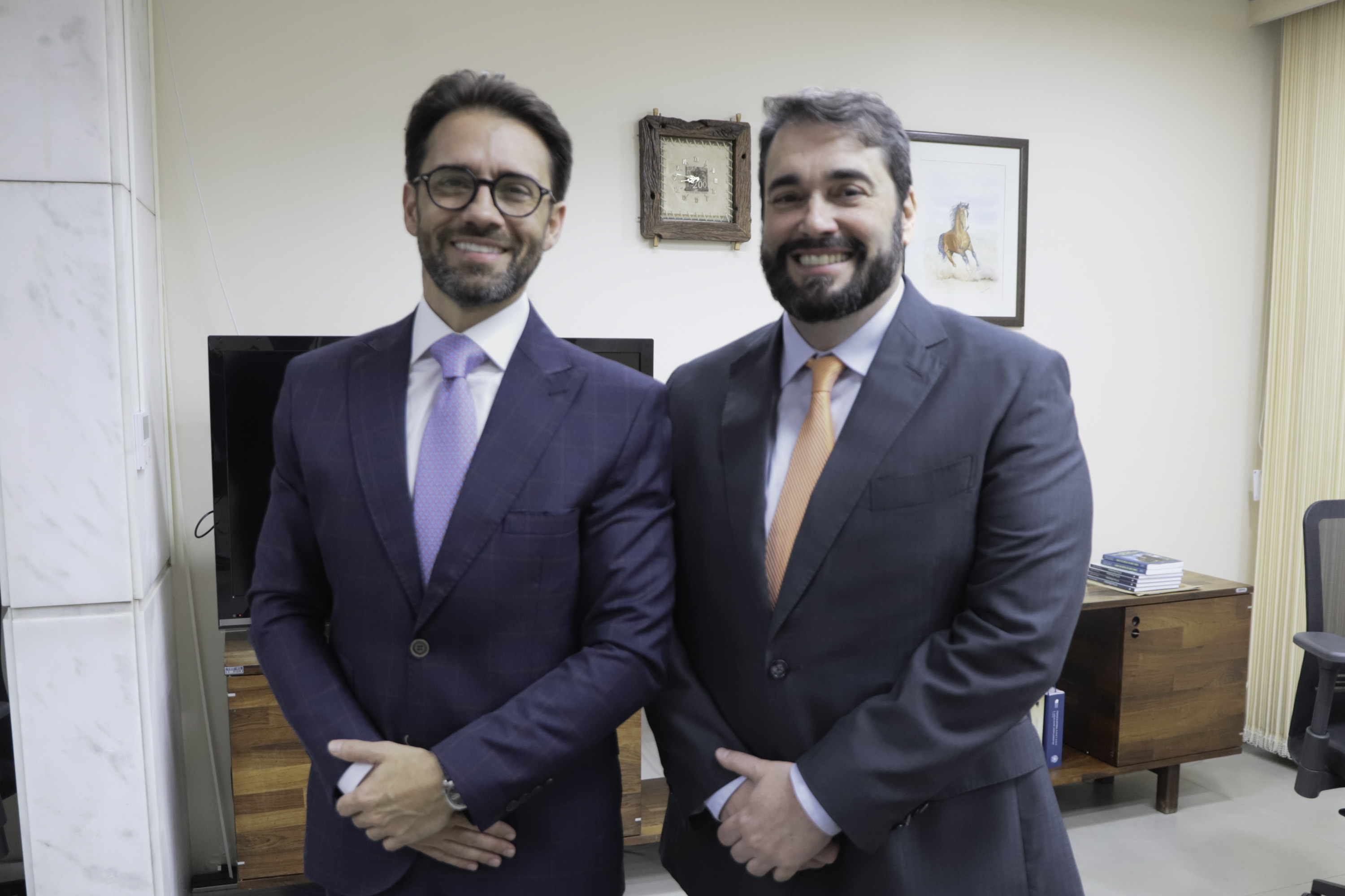 Marcelo e Alexandre posam sorrindo para a foto. Ao fundo aparecem quadros na parede. 
