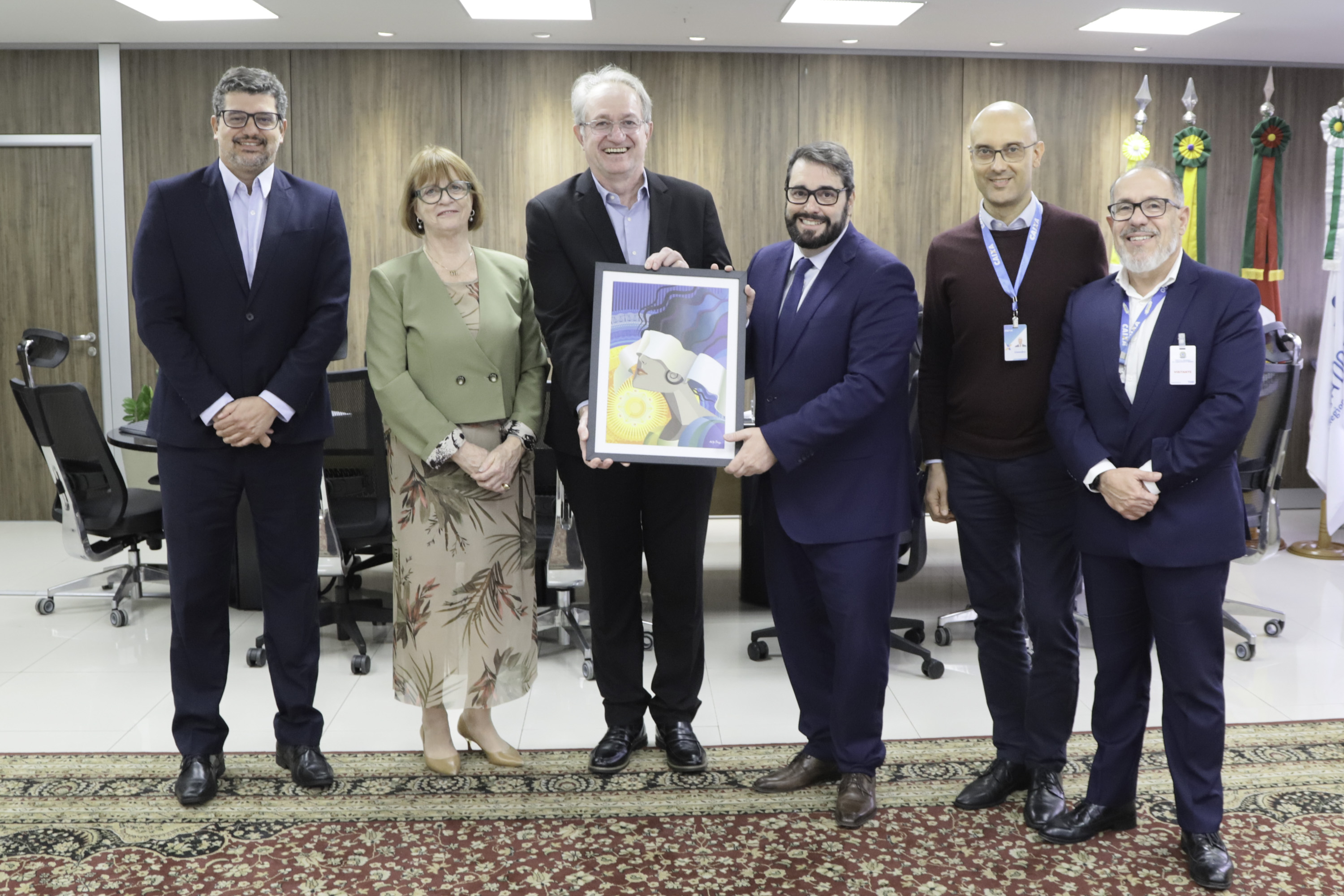 seis participantes da reunião posam de pé para a foto sobre um tapete. No centro, Renato e Alexandre seguram um quadro. Ao fundo aparecem bandeiras e a mesa de reuniões. 