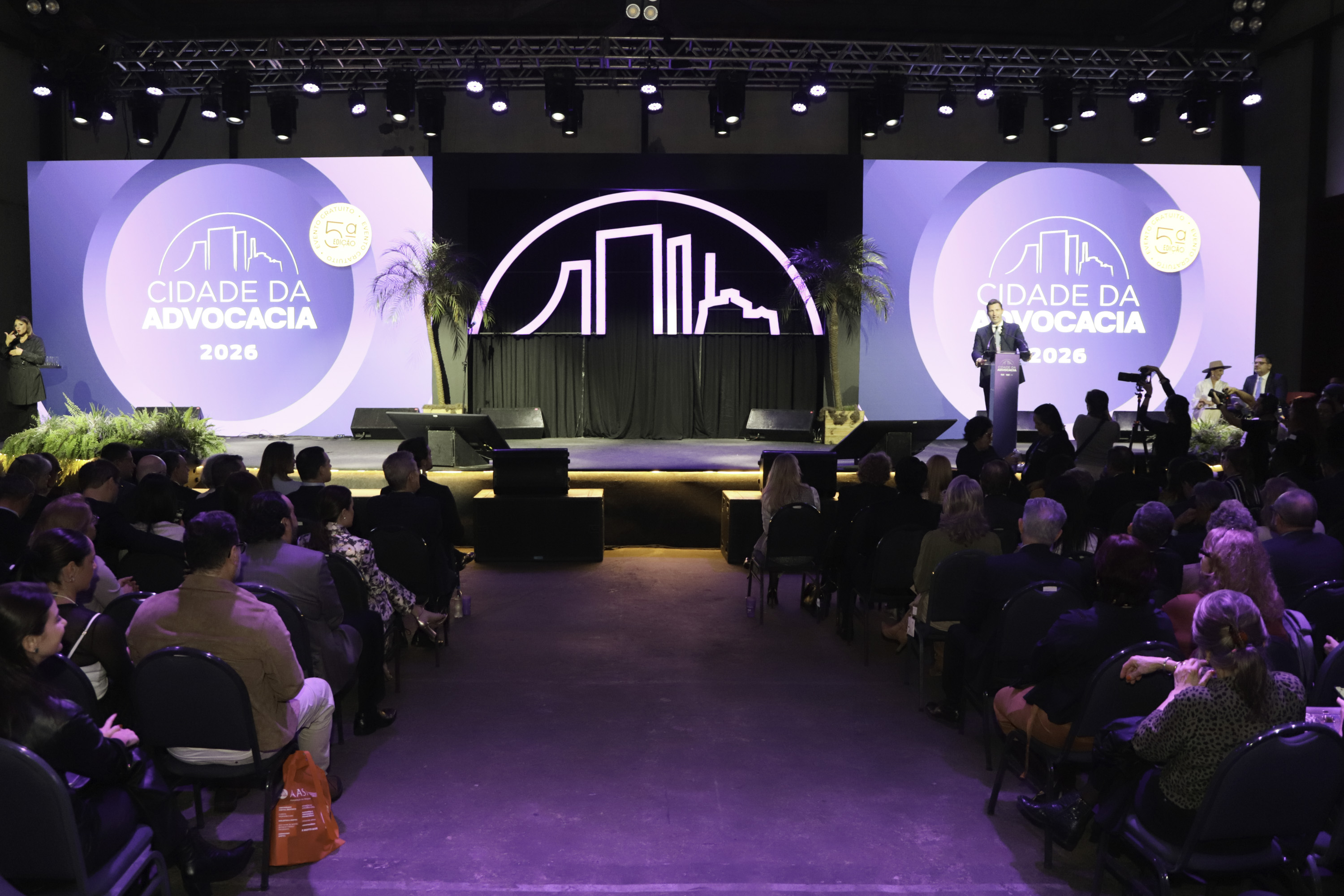 Público em primeiro plano e palco do evento ao fundo com alguém falando no púlpito