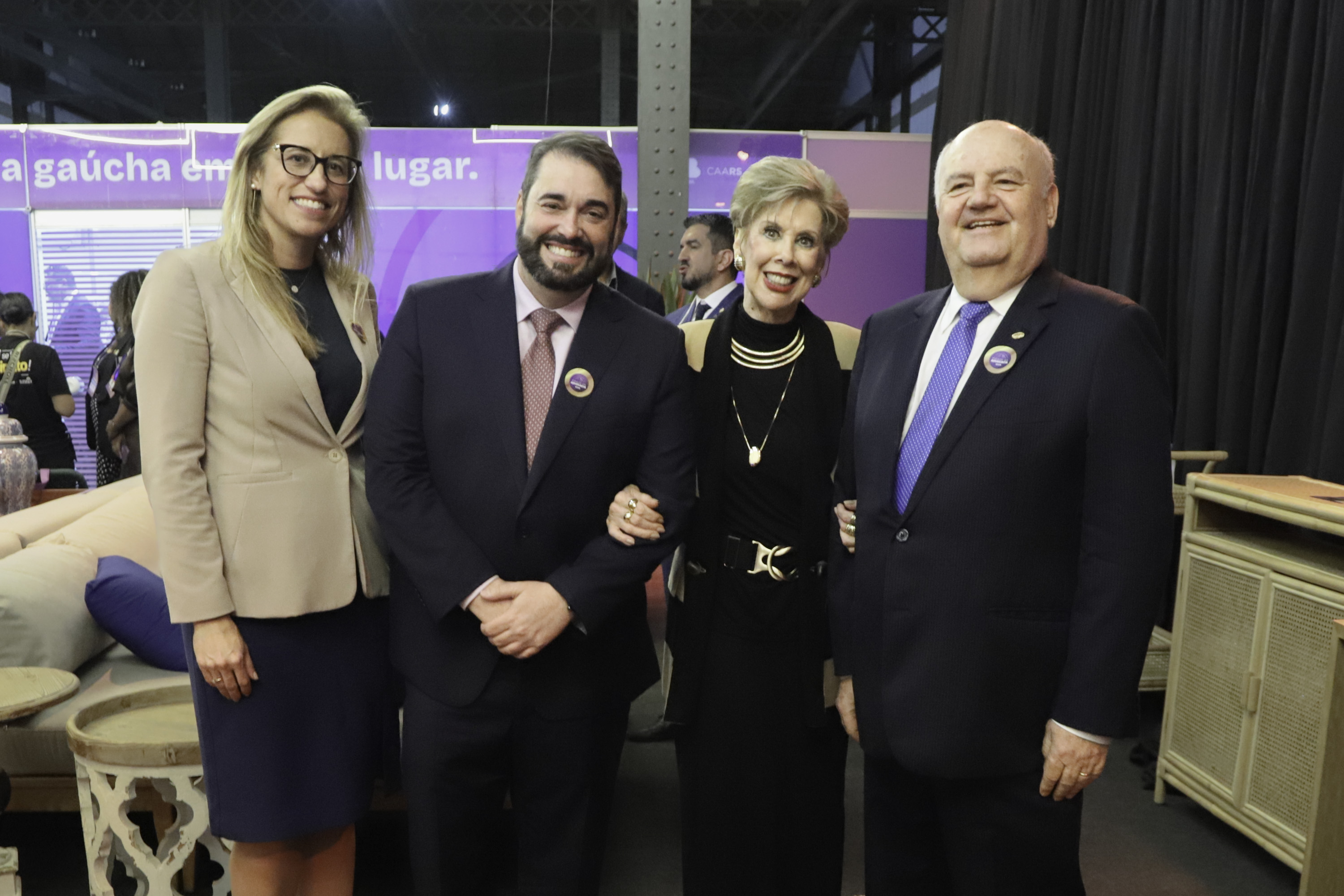 Grupo posa para a foto sorrindo, ao fundo aparece a movimentação de pessoas no evento. 