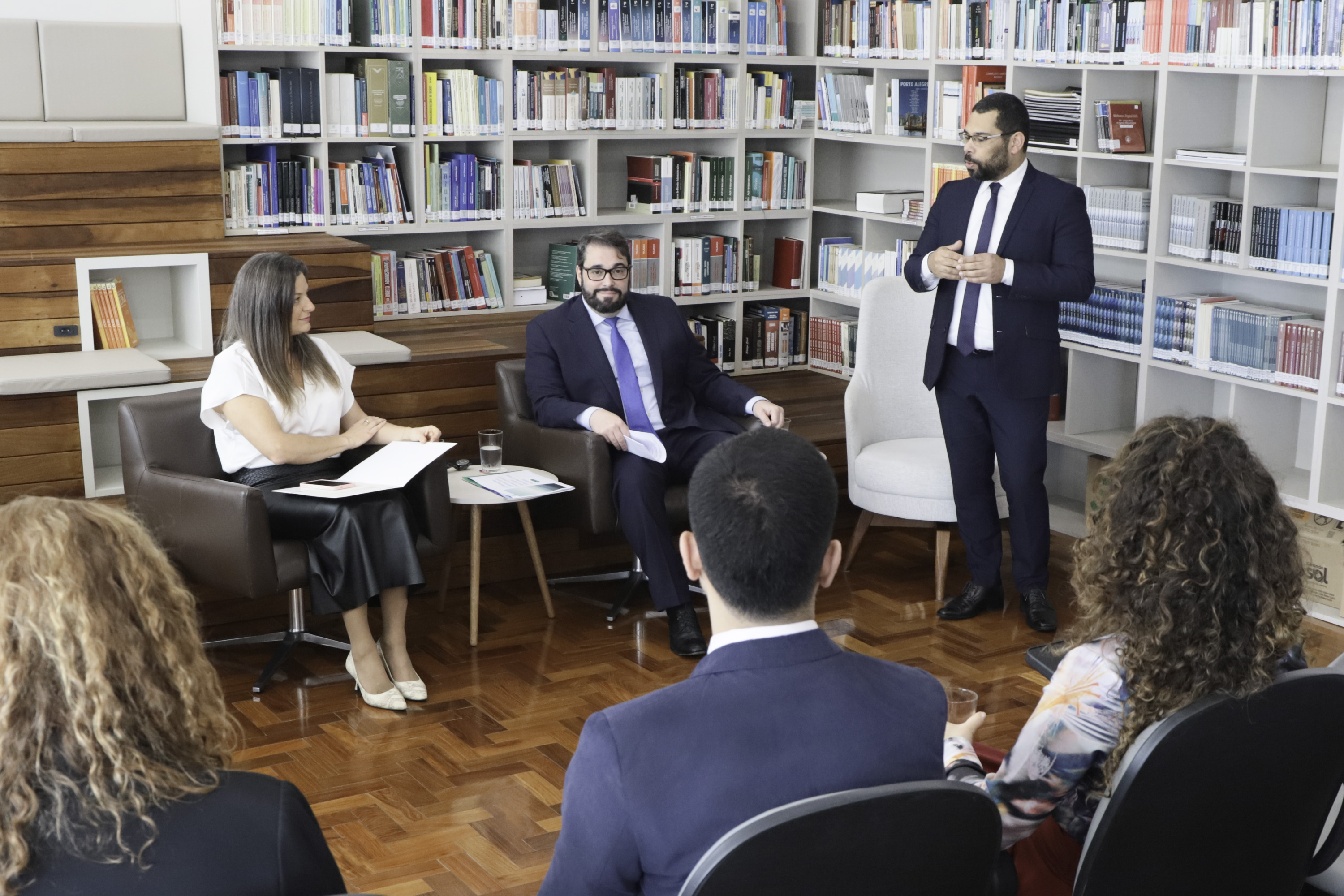 Os três aparecem ao centro, em frente à estrante de livros. Helena e Alexandre estão sentados em poltronas e Rafael fala de pé. 