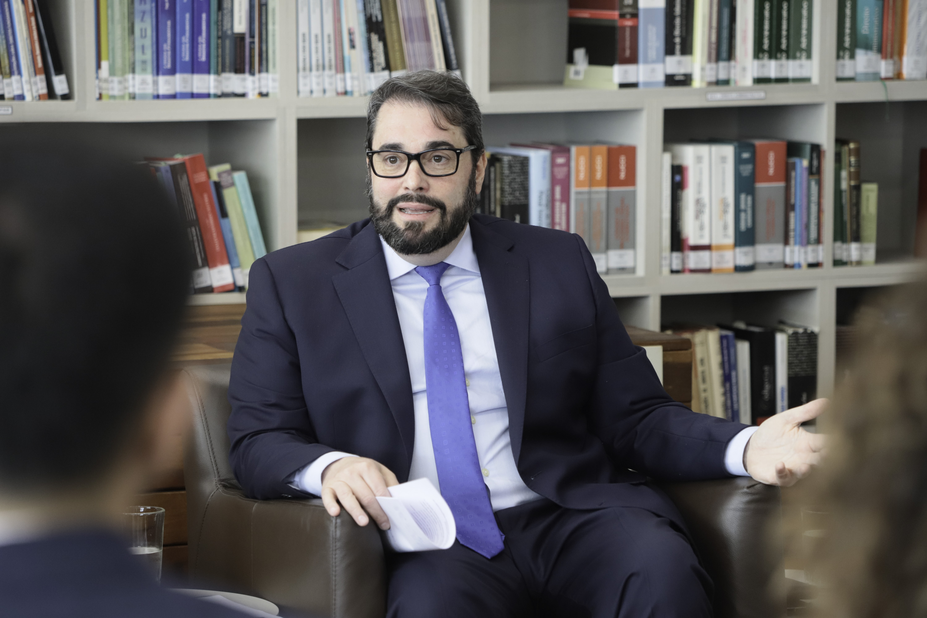 Alexandre é branco, tem cabelo e barba pretos escuros. Usa terno azul marinho. Está sendo em uma poltrona com uma estante ao fundo. Em primeiro plano, duas pessoas da plateia aparecem de costas, desfocadas.