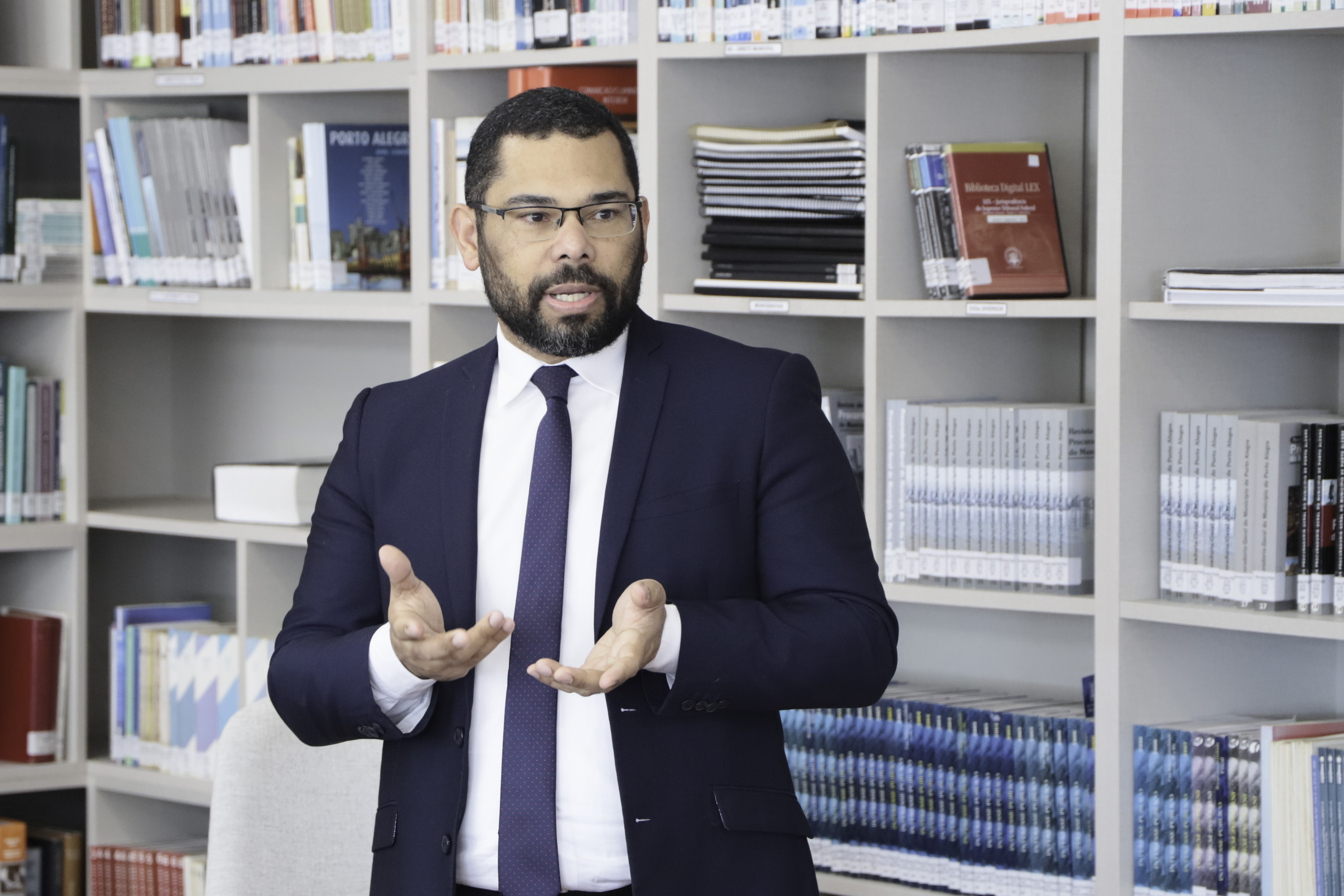 Rafael é negro, com cabelo e barba curtos escuro. Usa óculos e veste terno azul marinho. Fala de pé em frente à estante de livros. 