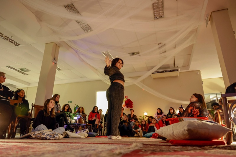 Imagem da roda do sarau com público em volta da slammer Mikaa, durante a sua apresentação. A artista está em pé de braços estendidos. Ela é uma mulher negra, veste um conjunto de moletom e calça pretos.