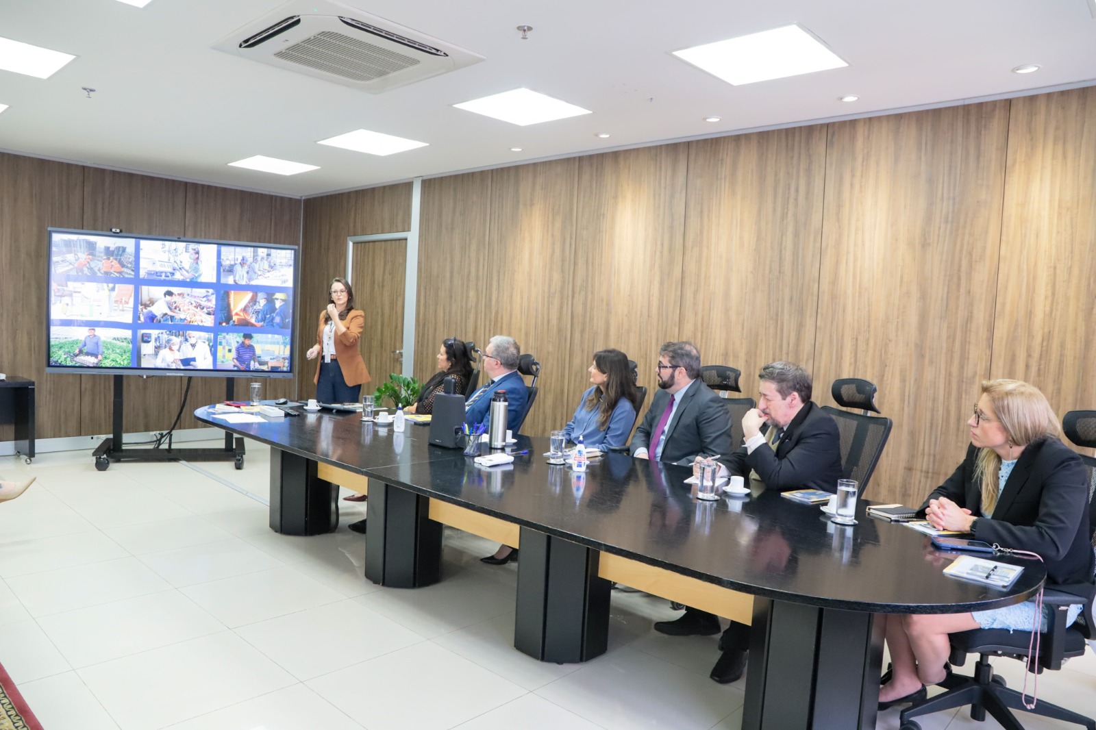 A foto mostra uma mulher fazendo uma apresentação numa tela grande de televisão com uma mesa de autoridades olhando.