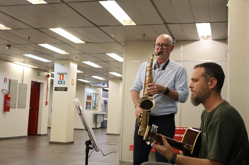 Imagem de Ricardo Hädrich e Rodrigo Hädrich. Ricardo está de pé tocando saxofone, ele é um homem de meia idade de pele clara, tem a cabeça raspada. Rodrigo está tocando violão sentado. Ele é um homem jovem, aparenta ter 30 anos, tem cabelos e barba castanhos, usa uma camiseta verde musgo.