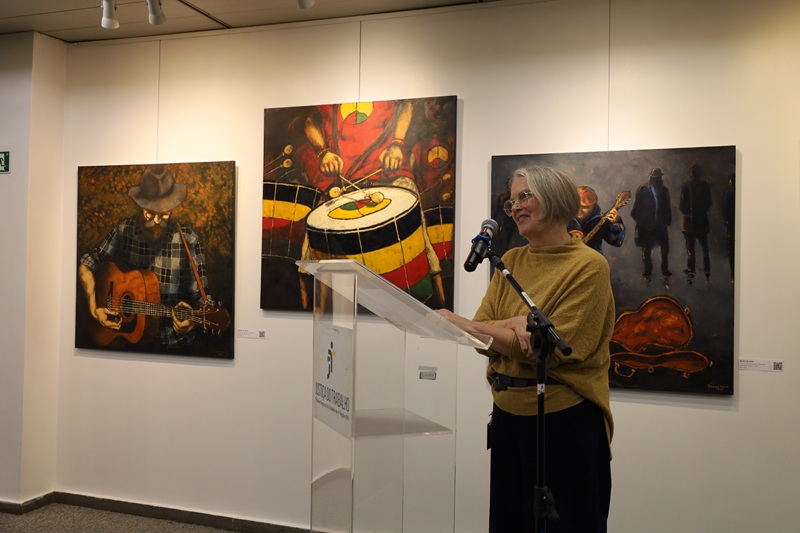 Imagem de Dagmar Ranck em pé falando ao microfone. Ela é uma mulher de meia idade, com pele clara e cabelos brancos curtos, usa óculos. Veste um pala bege e calças escuras.