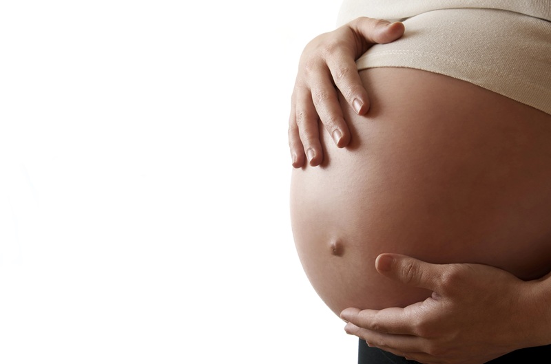 A fotografia mostra o ventre de uma mulher gestante, que tem as mãos ao redor da barriga.