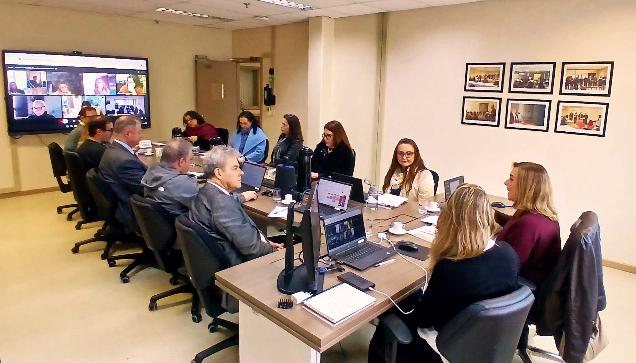 Pessoas participam de uma reunião em formato híbrido em uma sala bem iluminada. Homens e mulheres estão dispostos ao redor de uma mesa de reuniões com notebooks, blocos de anotações e xícaras de café. À esquerda, uma tela grande de televisão exibe a videochamada com diversos participantes remotos. Na parede bege, à direita, há seis quadros emoldurados com fotografias.