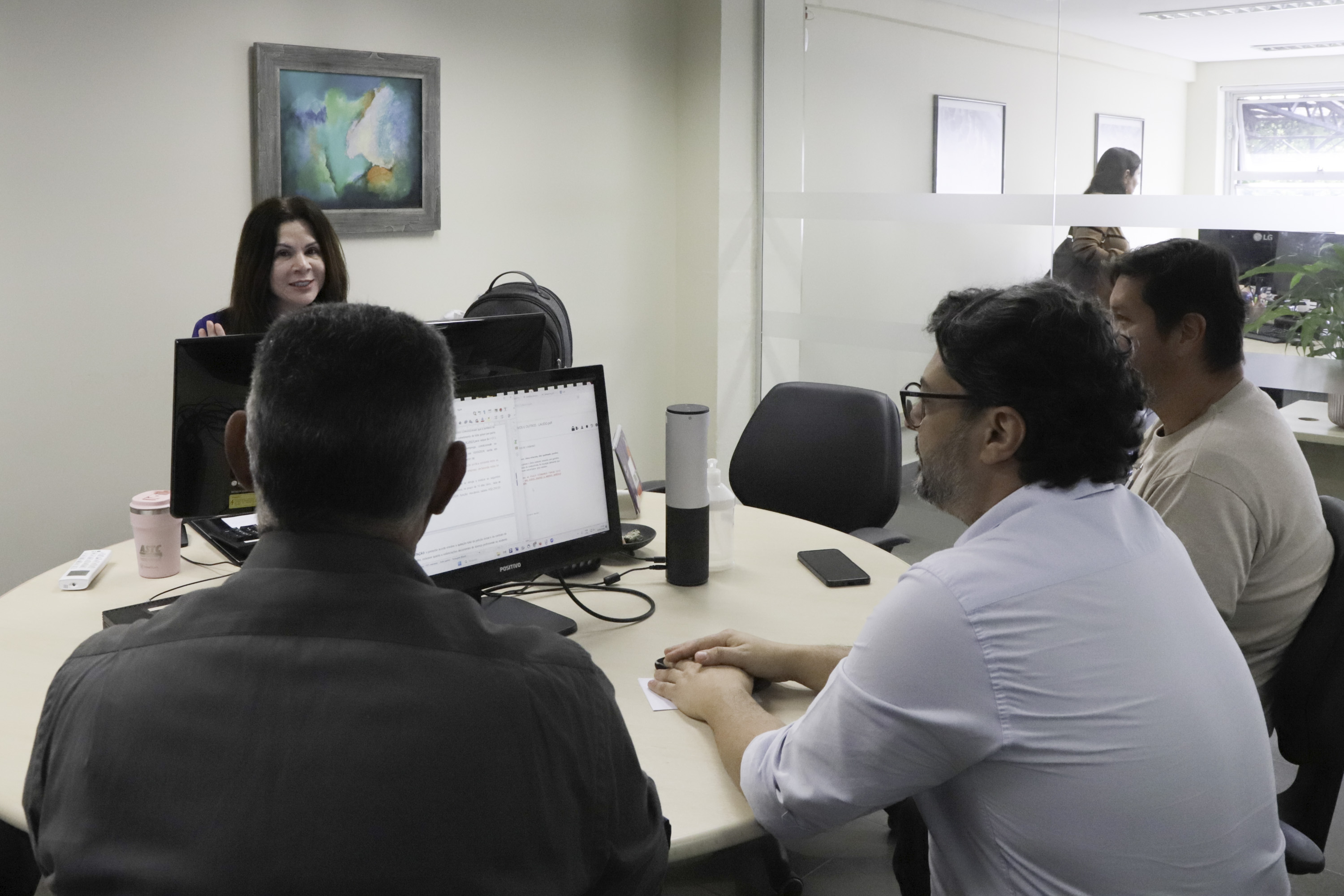 Servidora conduz audiência de conciliação. Ela aparece ao fundo. Em primeiro plano três pessoas de costas participam da audiência.