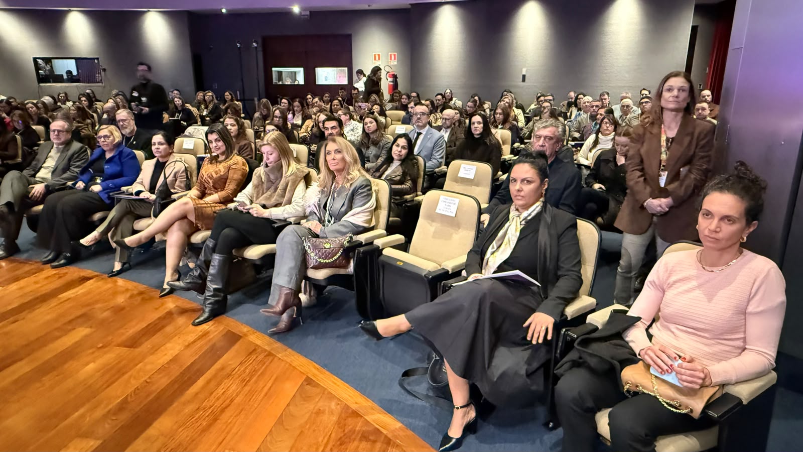 A foto mostra o público de um auditório com público grande sentado.