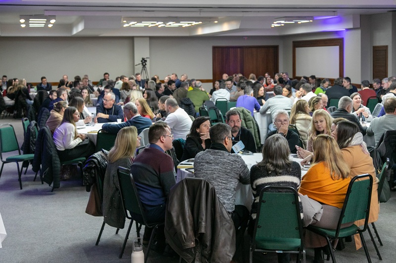 A fotografia mostra os participantes da edição do evento anterior reunidos em diversas mesas redondas, discutindo.