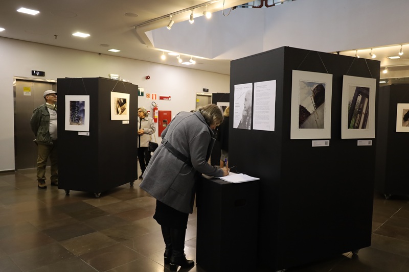 Imagem da exposição no saguão do tribunal. Na imagem aparece quatro painéis móveis onde as obras estão distribuídas, os painéis são pretos em formato de caixa retangular com rodinhas com quadros pendurados. Em frente ao painel mais próximo, está a lista de presença do evento em uma mesa em formato de caixa preta também, e uma mulher assinando. Em maior distância, um homem e uma mulher observam quadros pendurados em outro painel.