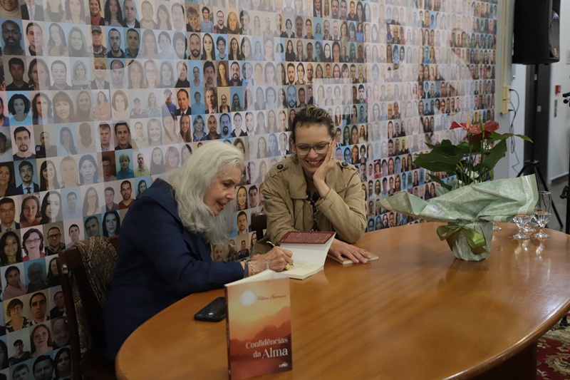 Imagem de Nilva autografando o livro de uma mulher. Nilva está sentada escrevendo e sorrindo levemente, a mulher ao lado dela sorri olhando para o livro com a mão apoiada no queixo.