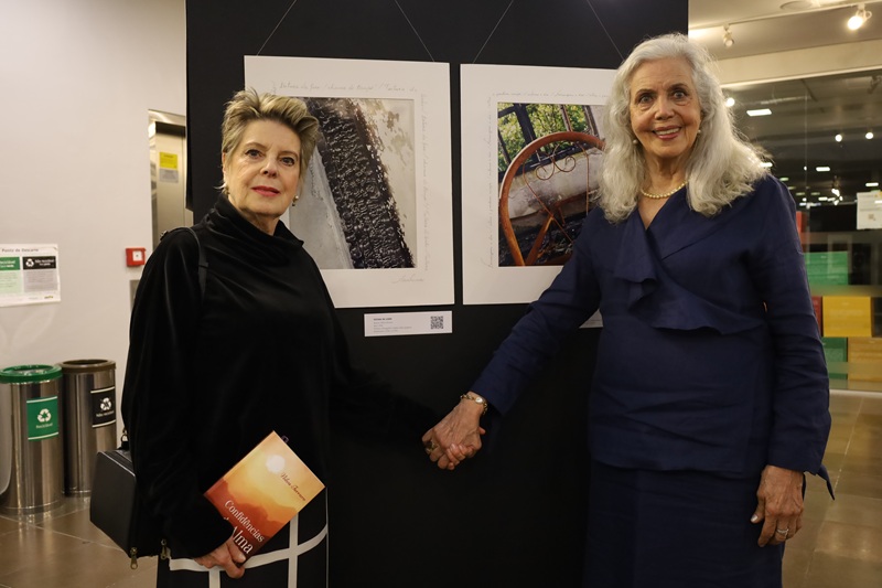 Imagem de Cristina Macedo e Nilva Ferraro de pé e de mão dadas em frente a duas obras. Cristina é uma mulher que aparenta ser de meia-idade, de pele clara, com cabelo curto, castanho com mechas loiras, ela veste um pala preto, bolsa preta e saia xadrez preto com listras brancas. Nilva é uma mulher que aparenta ser idosa, tem pele clara, cabelos brancos longos, veste um conjunto de saia e blusa azul escuro e um colar de pérolas.