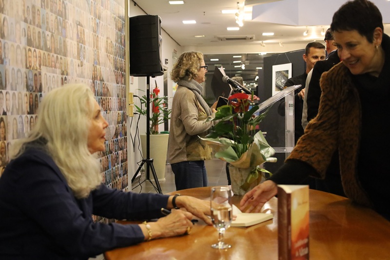 Imagem da sessão de autógrafos. À frente está a Nilva sentada olhando para uma mulher que estende o livro para autografá-lo. Atrás da mulher está uma fila de pessoas esperando autógrafo.