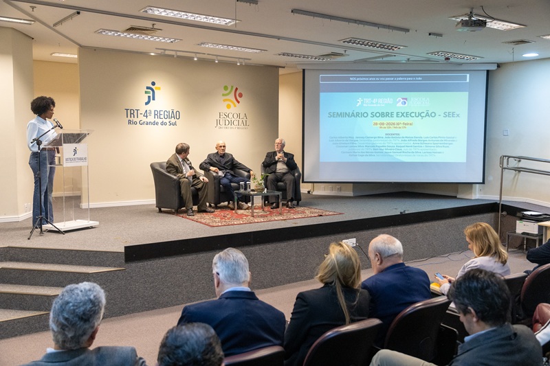 A fotografia mostra a mestre de cerimônias realizando a abertura do Seminário, no púlpito. Sentados em cadeiras, sobre o palco, estão os participantes da primeira palestra.