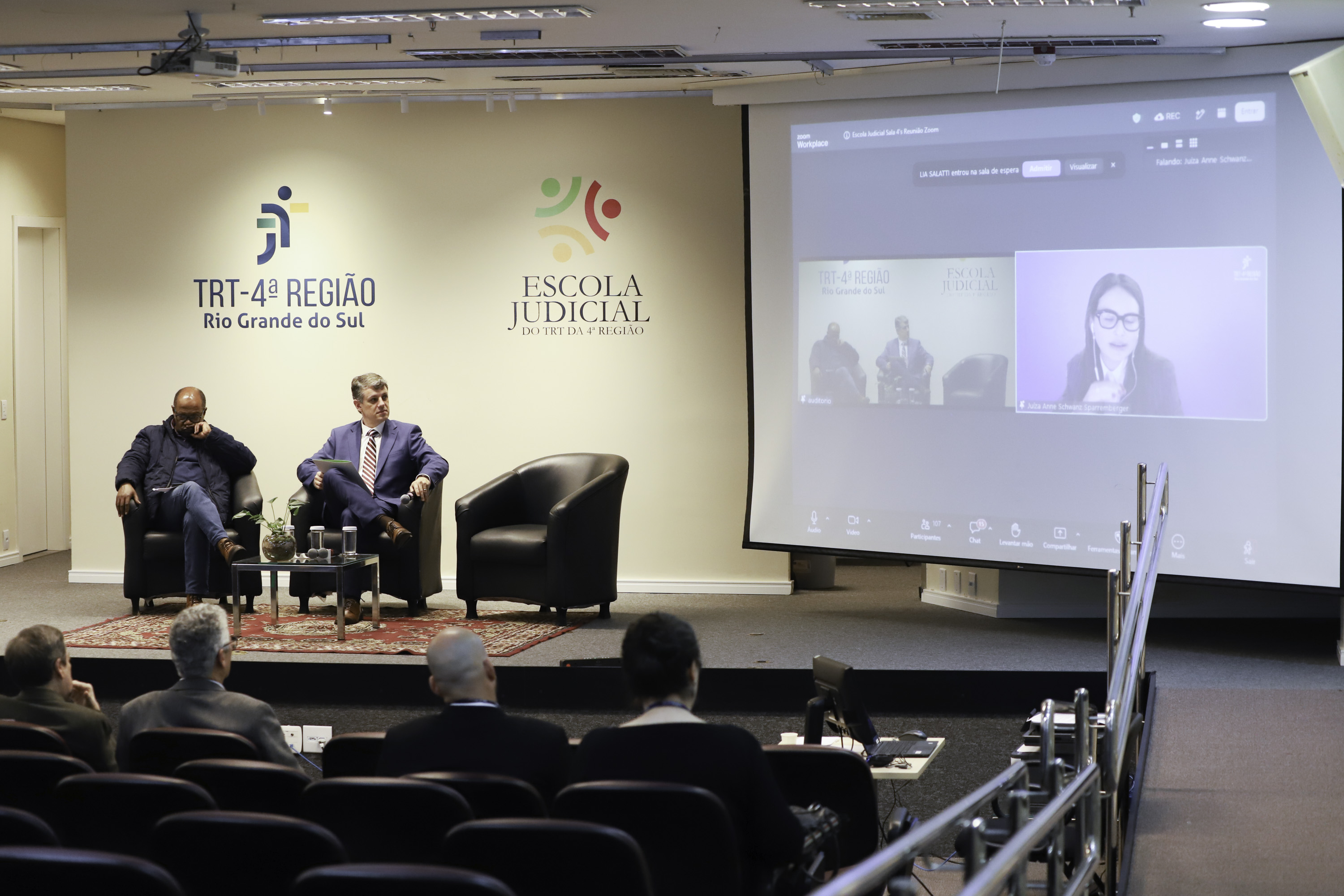 duas pessoas no palco e uma no telão à direita