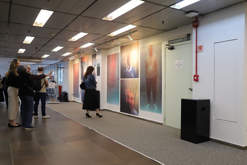 Imagem ampla da exposição. Na imagem, uma mulher está de costas em pé, ela observa uma das fotografias expostas na parede. A mulher aparenta ter cerca de 30 anos, veste uma saia longa preta, jaqueta jeans e salto alto preto. Atrás dela, um grupo de pessoas conversa de pé.