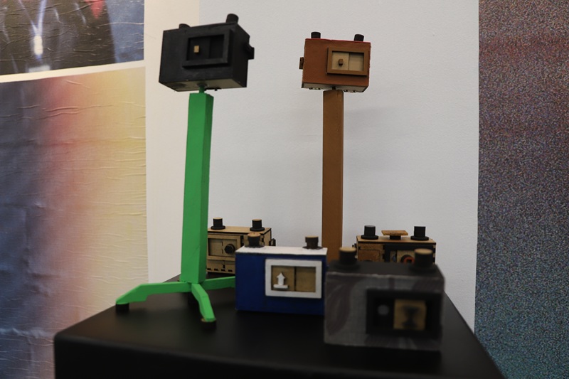 Imagem de seis câmeras pinhole em cima de um balcão preto. Duas das câmeras estão em cima de tripés feitos de peças de MDF, as câmeras também são todas de MDF. As câmeras e os tripés são pintados em diferentes cores: marrom, preto, azul, cinza e verde.