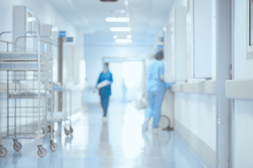 Trabalhadores em corredor de hospital. Imagem mostra duas pessoas usando uniformes azuis. O local é predominantemente branco. 