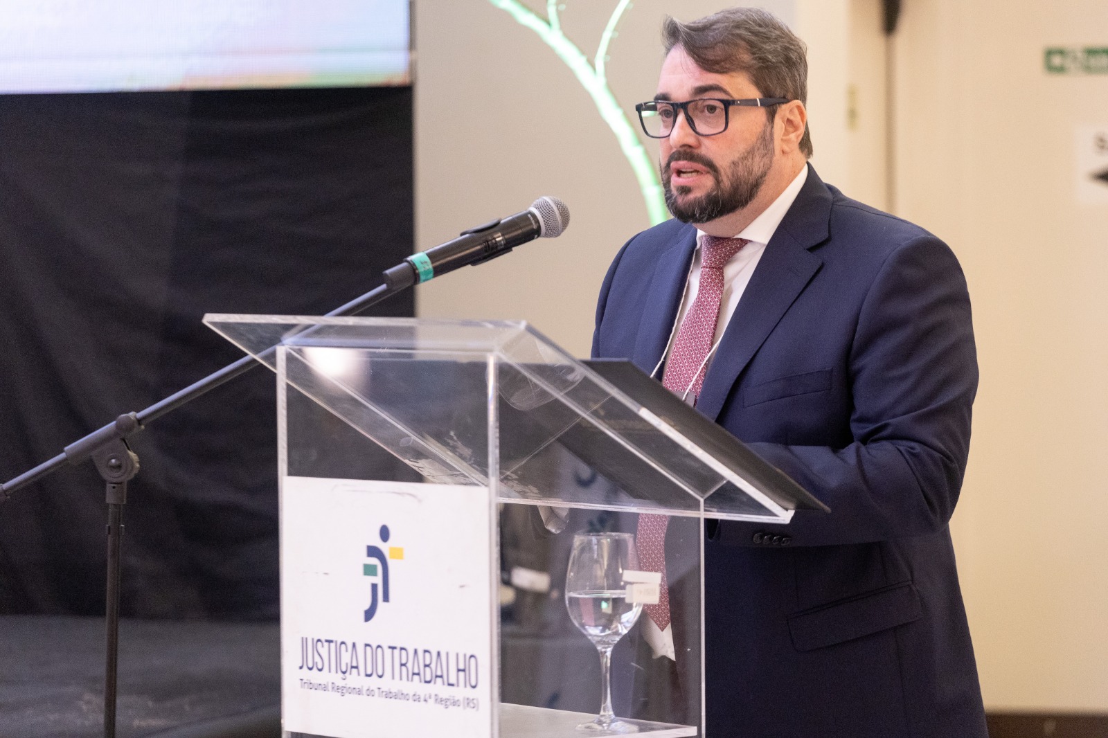 Alexandre é branco, usa cabelo e barba pretos, veste terno azul e gravata bordô. Fala no púlpito. 