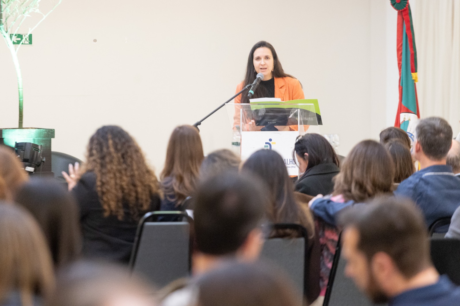 Caroline é branca, de cabelo liso preto Usa blazer laranha. Aparece falando no púlpito ao fundo, com o público de costas e desfocado no primeiro plano. 