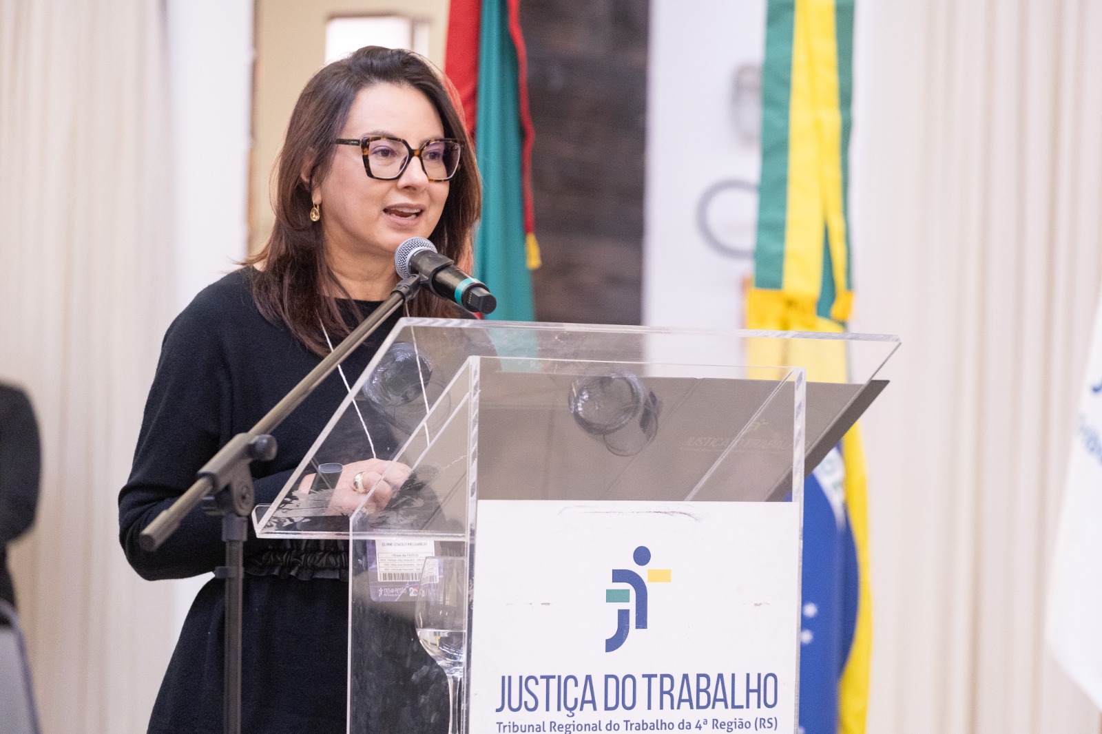 Eliane é branca, tem cabelo liso preto e usa óculos de aro grande. fala no púlpito. na frente dele aparece o logo da JT.