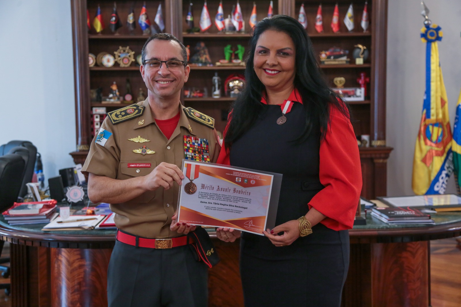 Tarciso e Tânia posam sorrindo para a foto. Eles seguram o certificado da homenagem. Ao fundo aparecem trofeuus, uma mesa de trabalho e bandeiras.