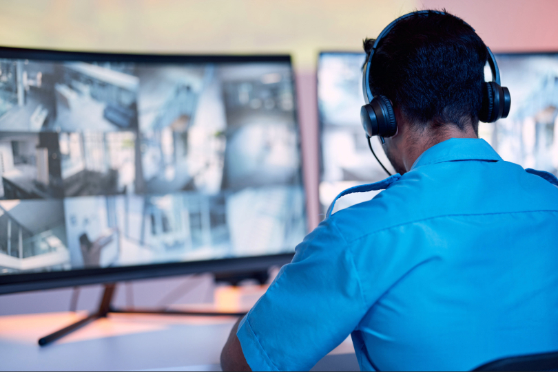 Homem faz vigilância por meio de dois monitores. Ele está de costas e usa uniforme azul.