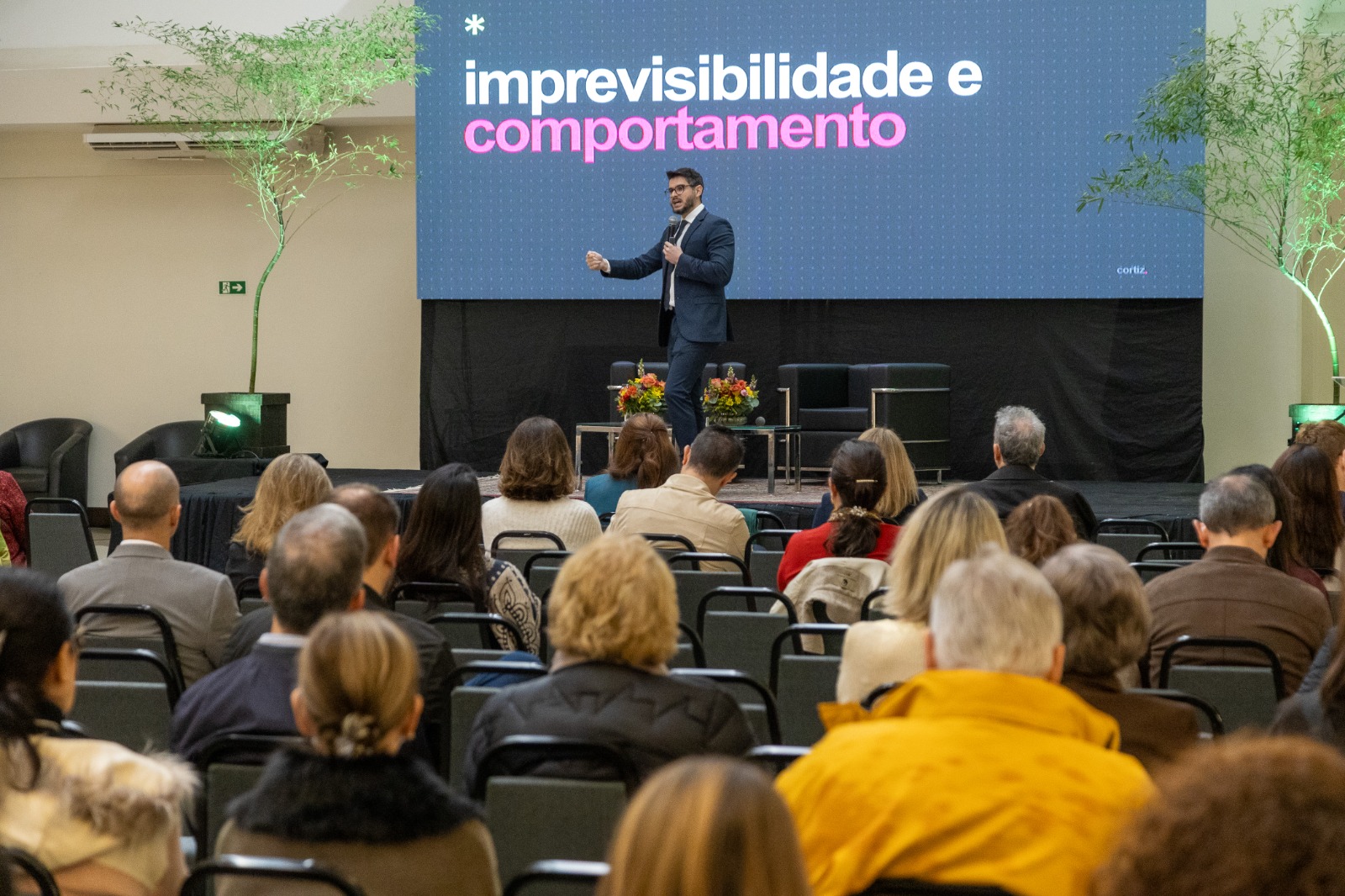 Foto em plano médio, mostrando parte do público e o palestrante ao fundo, no palco.;
