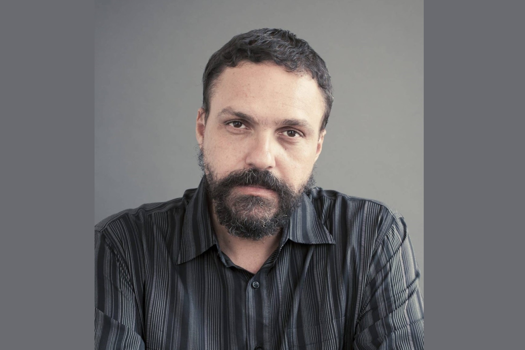 Paulo é pardo, tem cabelo e barba pretas e usa camisa cinza listrada.