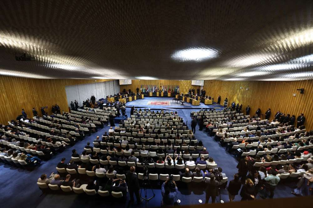 Foto tirada de um ângulo alto do Plenário, mostrando as pessoas assistindo e outras nas mesas.