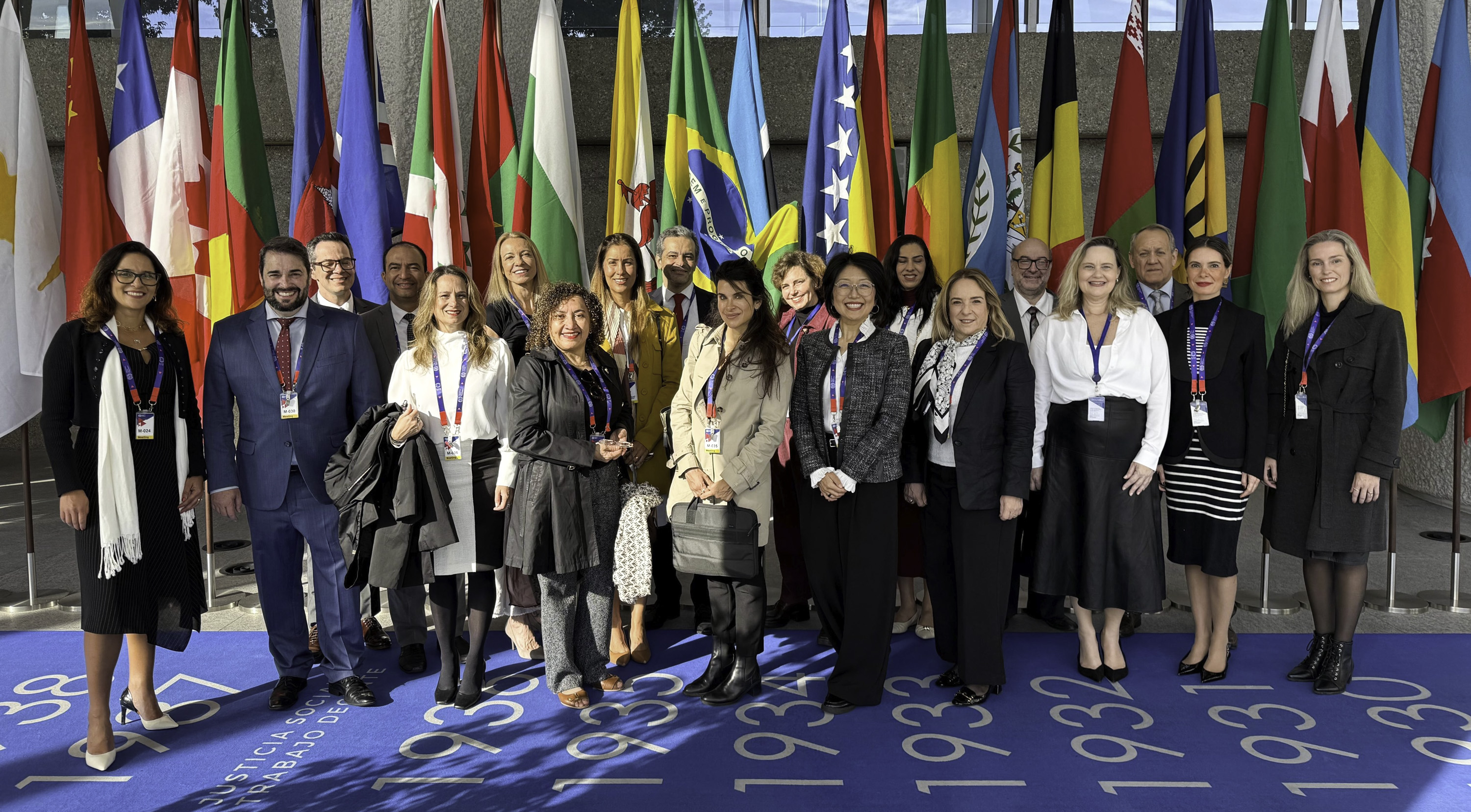 23 pessoas posam para a foto sorrindo. ao fundo aparecem bandeiras diversos países.