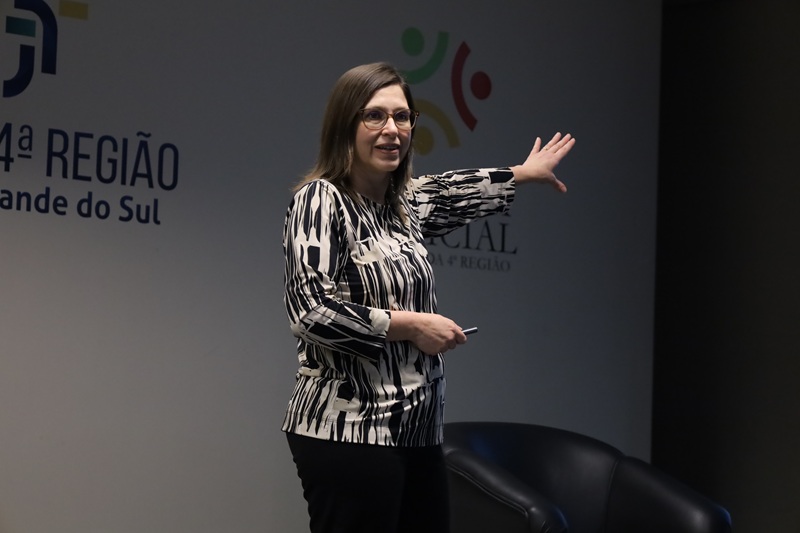 Imagem da palestrante Bibiana em pé no palco do auditório. Ela é uma mulher de meia idade, pele clara, cabelos lisos castanhos. Veste uma blusa branca estampada em preto e uma calça preta. Ela usa óculos e está estendendo a mão esquerda em direção ao projetor.