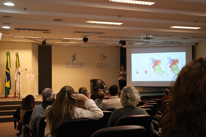 Imagem da plateia em primeiro plano, ao fundo está o palco com a palestrante apresentando.