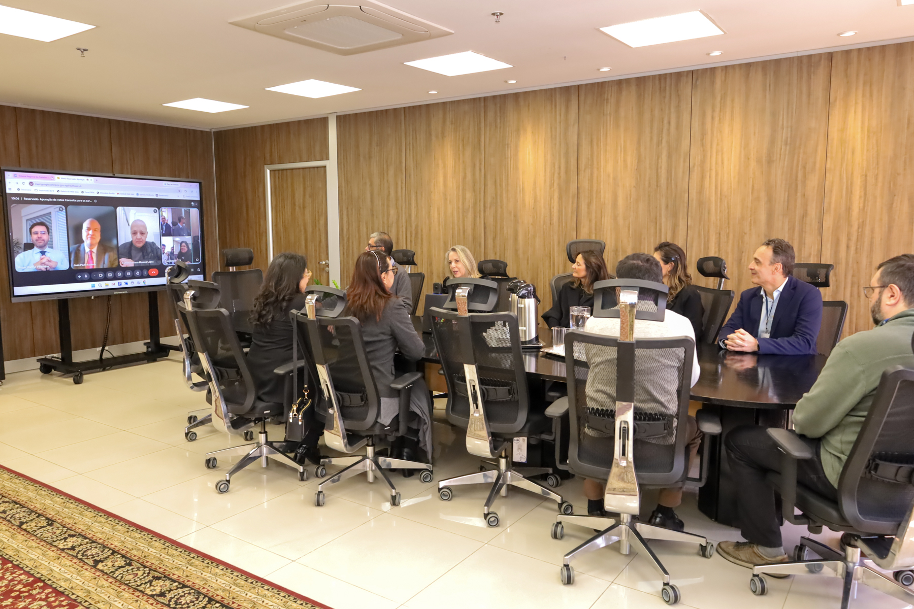 Foto em ângulo aberto, mostrando os participantes da reunião sentados à mesa do Salão Nobre e o telão no fundo, aparecendo quem participou por vídeo. Caption