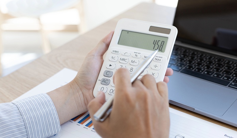 A fotografia mostra em close as mãos de trabalhador usando uma calculadora e uma caneta.