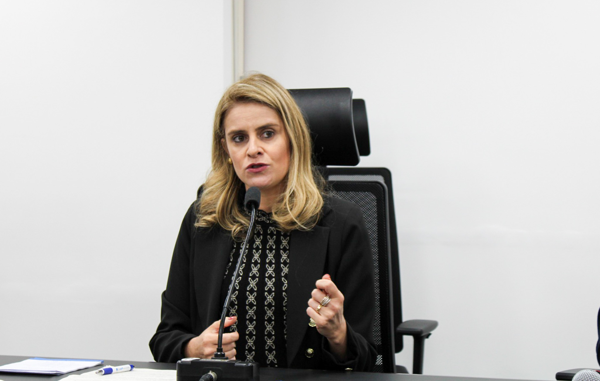 A foto mostra uma mulher loira sentada, falando ao microfone.