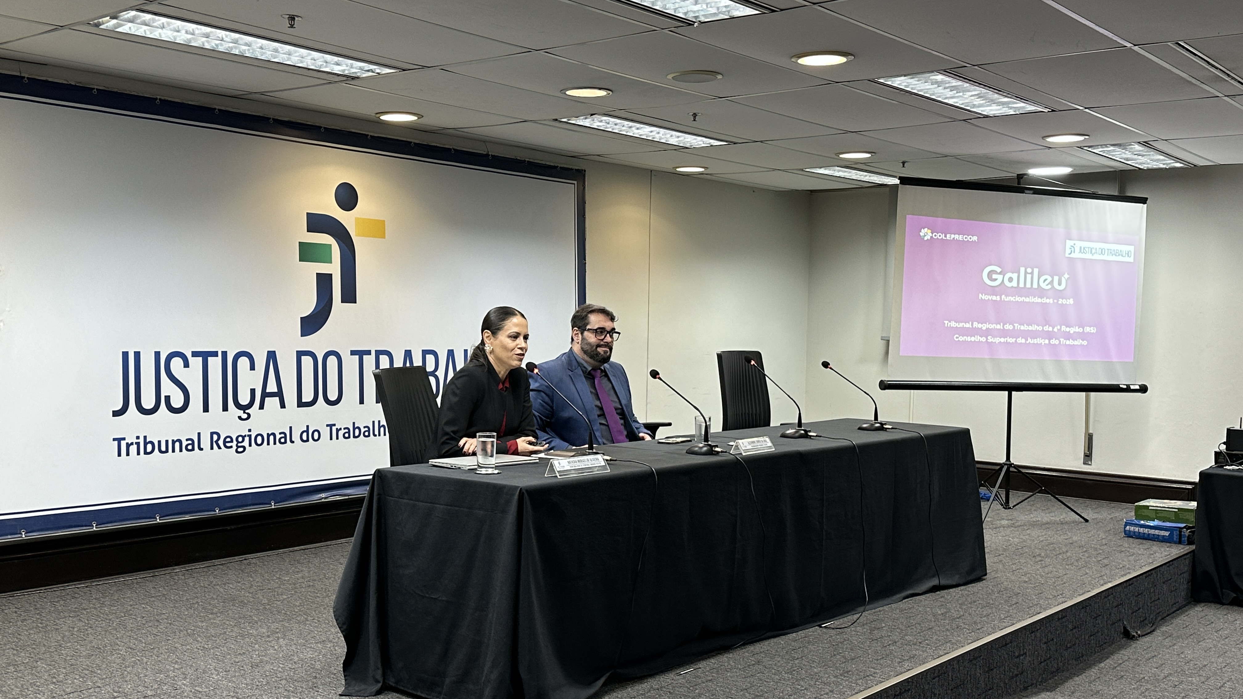 A foto mostra duas pessoas sentadas com painel ao lado fazendo uma apresentação.