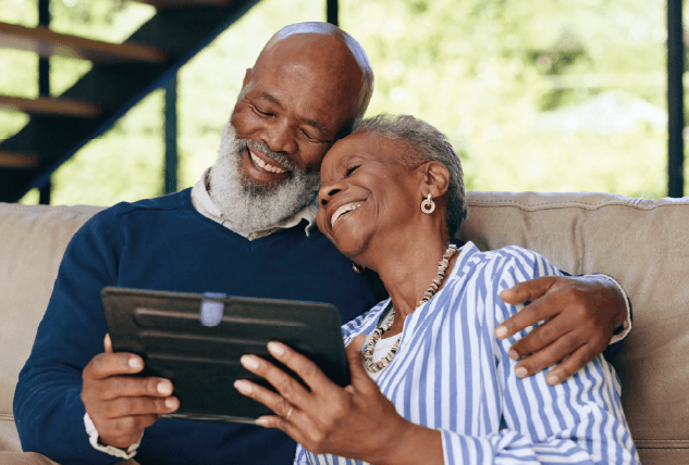 Um casal sénior negro partilha um momento de carinho e cumplicidade num sofá confortável. O homem, de barba e cabelo grisalhos, veste uma camisola azul e abraça a mulher pelos ombros. Ela, vestida com uma camisa às riscas azuis e brancas, apoia a cabeça no ombro dele enquanto ambos sorriem alegremente para o ecrã de um tablet que seguram juntos. A imagem transmite afeto, aconchego e felicidade num ambiente doméstico e luminoso.