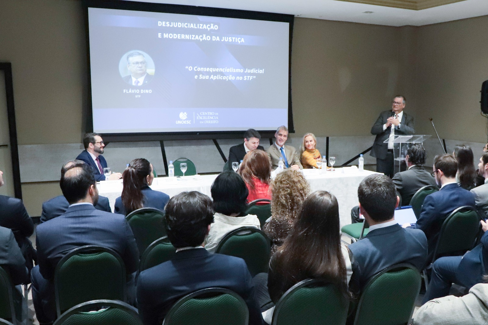 Foto geral do evento, com mesa oficial e palestrantes ao fundo e público de costas em primeiro plano