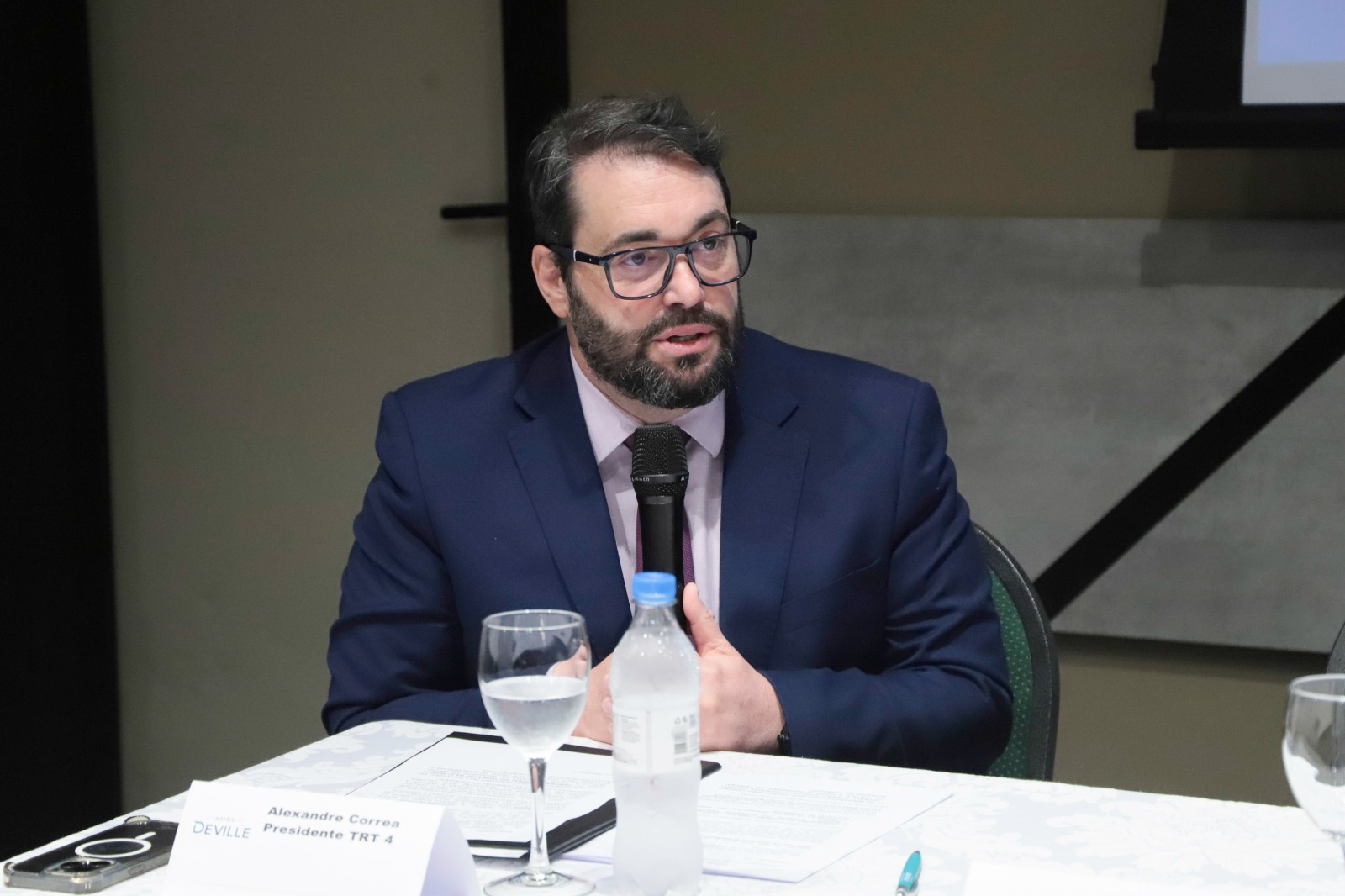 Alexandre é branco, tem cabelo e barba escuras e usa ólculo e armação garnde. Fala na mesa oficial.